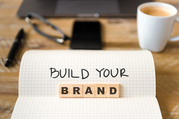 Closeup on notebook over vintage desk background, front focus on wooden blocks with letters making Build Your Brand text