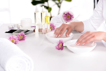 Obraz na płótnie Canvas cropped image of woman receiving bath for nails at table with flowers, towels, candles, aroma oil bottles, nail polishes, cream container and tools for manicure in beauty salon