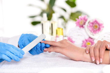 Obraz na płótnie Canvas partial view of beautician in latex gloves doing manicure by nail file to woman at table in beauty salon