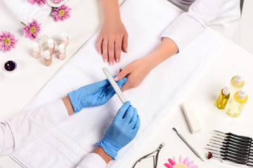 elevated view of beautician in latex gloves doing manicure to woman by nail file at table with flowers, towels, nail clippers, candles, nail polishes, aroma oil bottles and cuticle pusher