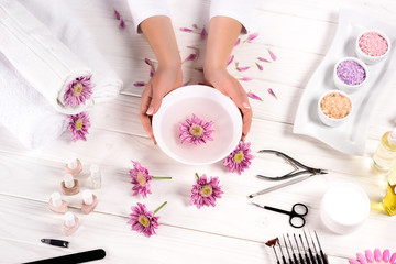 partial image of woman holding bath for nails over table with flowers, towels, colorful sea salt, aroma oil bottles, nail polishes, cream container and tools for manicure in beauty salon