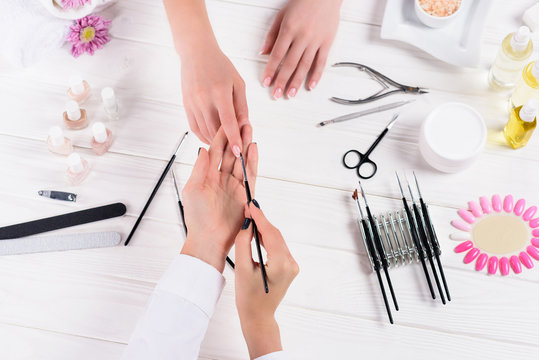 cropped image of beautician doing manicure to woman at table with nail polishes, nail files, nail clippers, cuticle pusher, sea salt, flowers, aroma oil bottles and samples of nail varnishes