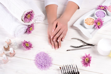 Fototapeta na wymiar cropped shot of female hands at table with towels, flowers, nail polishes, colorful sea salt, cream container and tools for manicure in beauty salon