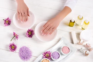 Obraz na płótnie Canvas cropped image of woman receiving bath for nails at table with flowers, colorful sea salt, cream container, aroma oil bottles and nail polishes in beauty salon