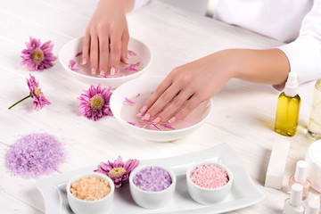 Obraz na płótnie Canvas partial view of woman receiving bath for nails at table with flowers, colorful sea salt, cream container, aroma oil bottles and nail polishes in beauty salon