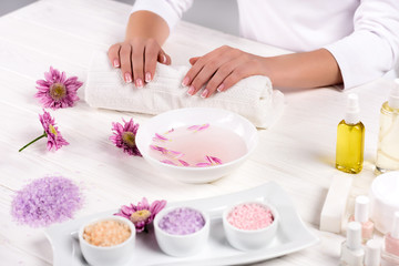 Fototapeta na wymiar cropped image of woman holding hands on towel for manicure procedure at table with flowers, colorful sea salt, cream container, aroma oil bottles and nail polishes in beauty salon