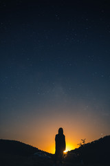 alone woman silhouette in mountains, girl wrapped up in a plaid looks at the sky, enjoying view of amazing night sky full of stars, vertical photo