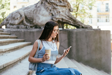 young woman in eyeglasses holding paper cup and using smartphone while sitting on stairs