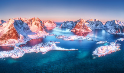 Aerial view of Reine and Hamnoy at sunrise in winter. Lofoten islands, Norway. Panoramic landscape with blue sea, snowy mountains, high rocks, village, buildings, rorbuer, sky in the morning. Top view