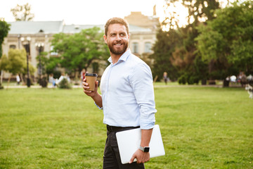 Image of successful business man in white shirt, walking through park carrying takeaway coffee and...