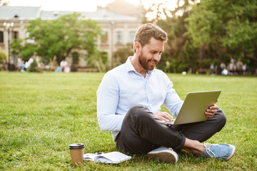 Photo of businesslike pleased man in business clothing, sitting on grass in park with legs crossed and working on silver laptop - obrazy, fototapety, plakaty
