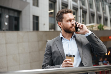 Photo of mature man in gray suit looking aside, while standing and holding takeaway coffee in front of modern business center during business call