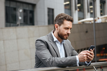 Photo closeup of adult businesslike man in gray suit typing or reading text message in cell phone, while standing in front of modern office building