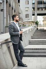 Full length profile picture of confident businessman 40s in gray suit, standing and drinking takeaway coffee in front of modern business center