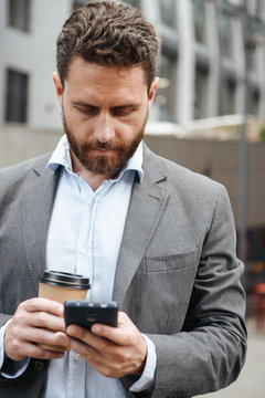 Photo of adult man in gray suit typing or reading text message in mobile phone, while standing with takeaway coffee in front of modern business center