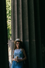 beautiful smiling young woman in hat holding coffee to go and standing between columns