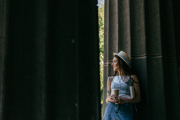 beautiful smiling young woman with book and coffee to go standing between columns and looking away