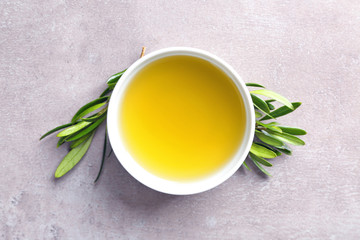 Bowl with olive oil and green leaves on grey background, top view