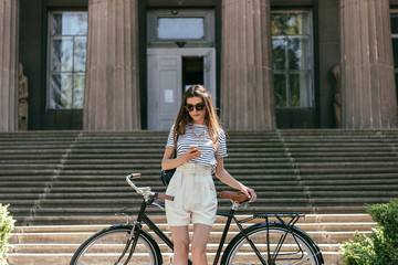 Obraz na płótnie Canvas attractive girl in sunglasses using smartphone while standing with bike near beautiful building with columns and stairs