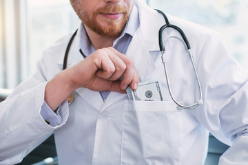 Good day. Satisfied bearded doctor wearing a uniform and putting money into his pocket