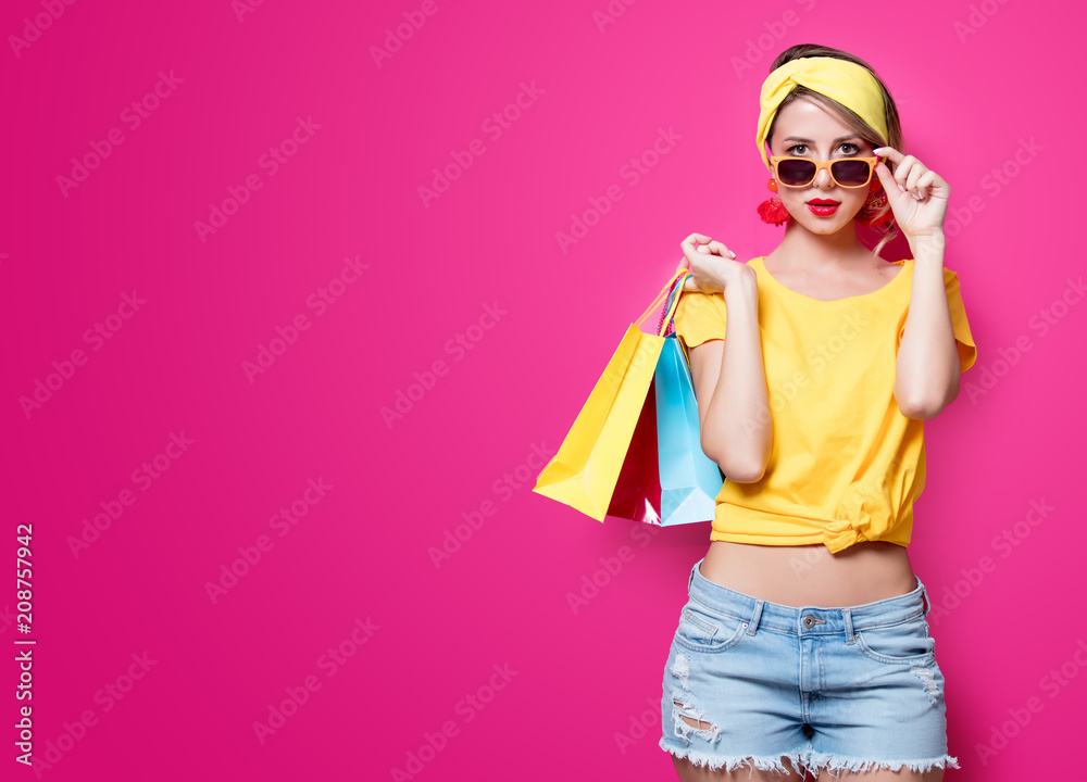 Wall mural Young redhead girl in yellow t-shirt and blue jeans holding a colorful bags on pink background