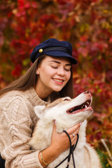 portrait of young beautiful woman in hat sitting in autumn park hugging white husky dog. happiness and friendship. girl playing with pet on red trees background