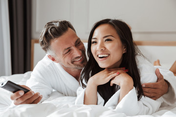 Photo of multiethnic couple caucasian man and asian woman wearing white bathrobe lying in home...