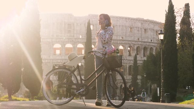Beautiful young woman in colorful fashion dress with bike talking on phone call in front of colosseum in Rome at sunset with trees happy attractive tourist girl in colle oppio