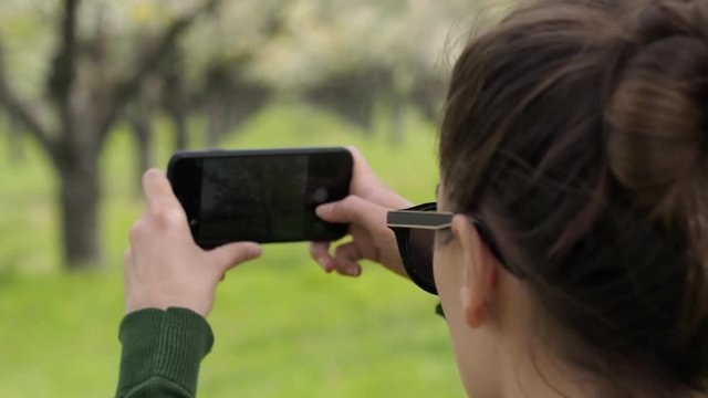 Rear view of a young caucasian woman in the nature taking photos with her smartphone