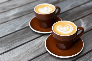 Two dark red cups of cappuccino with the heart shape latte-art on wood background