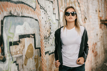 Girl wearing t-shirt, glasses and leather jacket posing against street , urban clothing style. Street photography