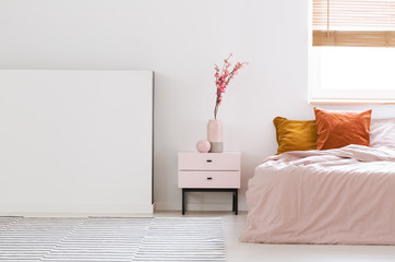 Real photo of a pink, feminine bedroom interior with orange cushions on bed and flowers on a nightstand next to an empty wall - obrazy, fototapety, plakaty
