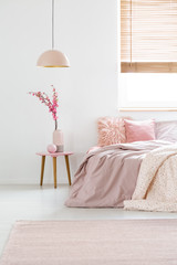 Real photo of flowers on a bedside table, peach lamp and pink bed in simple, pastel bedroom interior