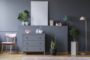 Pink chair next to grey cabinet in living room interior with plants and mockup of poster. Real photo