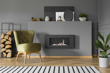 Green armchair and firewood next to fireplace in grey living room interior with plant. Real photo