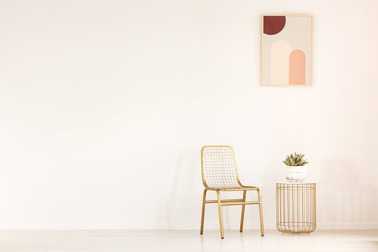 Real photo of a gold chair standing next to a small table with a plant in minimal living room interior with a poster on a wall