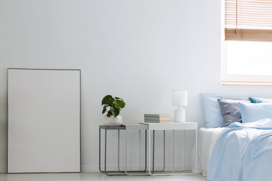 Real photo of an empty poster and metal tables standing against white wall in bright bedroom interior with blue bed and window. Paste your graphic here