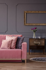 Cropped photo of a sofa, pillows, cabinet with flowers and empty golden frame on a grey wall