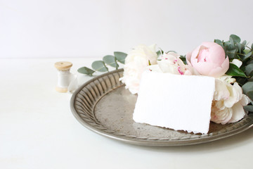 Feminine wedding or birthday table composition with floral bouquet. White and pink peonies flowers and eucalyptus branches on old vintage silver tray, craft paper card and silk ribbon.