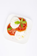 Mozzarella and tomatoes with basil on a white square plate. The background is white. Vertically. Copiespice.