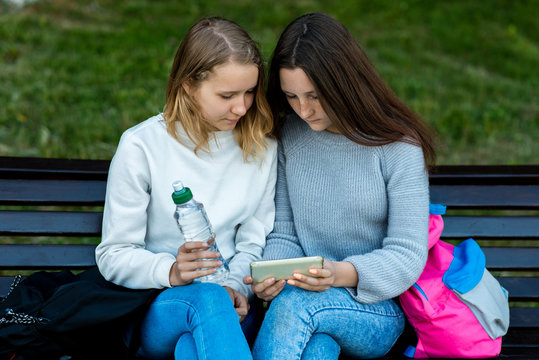 Two Girlfriends Student. Summer In Nature. They Sit On A Bench Watching A Movie In Smartphone. Holds His Hands A Bottle Of Water. Watching The Video On The Phone. Rest After School.