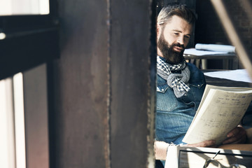 Male writer with dark hair and beard, weared in denim jacket and scarf is sitting in the modern office near the window and reading his notes