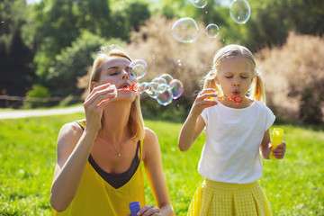 Relaxing outdoors. Beautiful caring mother blowing soap bubbles with her daughter while having fun in the park