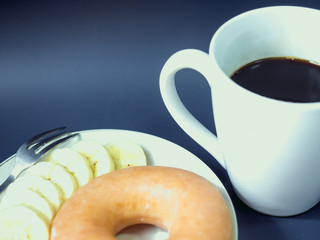 close up from banana and donut with black coffee in white cup from morning time with blue background