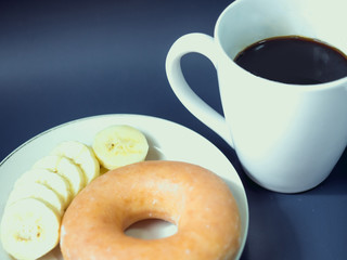 close up from black coffee in white coffee cup with soft focus (banana and donut) at foreground put on blue background