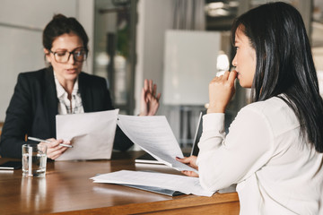 Fototapeta na wymiar Image of nervous asian woman looking and talking to businesswoman, while sitting at table in office during job interview - business, career and recruitment concept