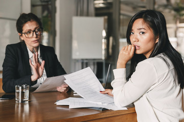 Uptight asian woman expressing fear and uncertainty while sitting at table in office and talking to female employee, during job interview - business, career and recruitment concept