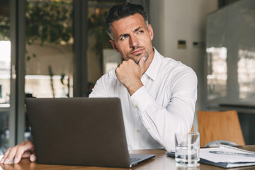 Fototapeta na wymiar Photo of european man wearing white shirt and earbud sitting at table in office with brooding look aside, while working on laptop
