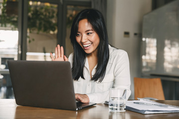 Image of happy asian woman 20s wearing white shirt smiling and waving hand at laptop, while speaking or chatting on video call in office