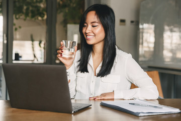 Joyful asian office woman 20s wearing white shirt smiling, while looking at screen of laptop and drinking water from glass